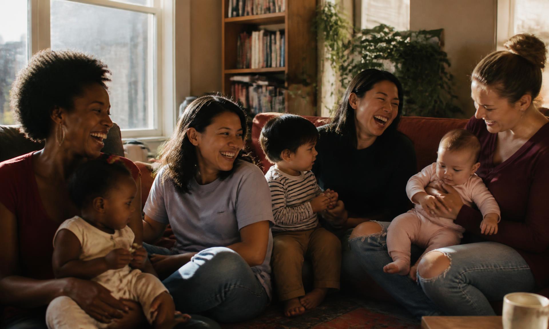 Four mothers of different backgrounds laughing together on a sofa with their children in a warm sunlit living room